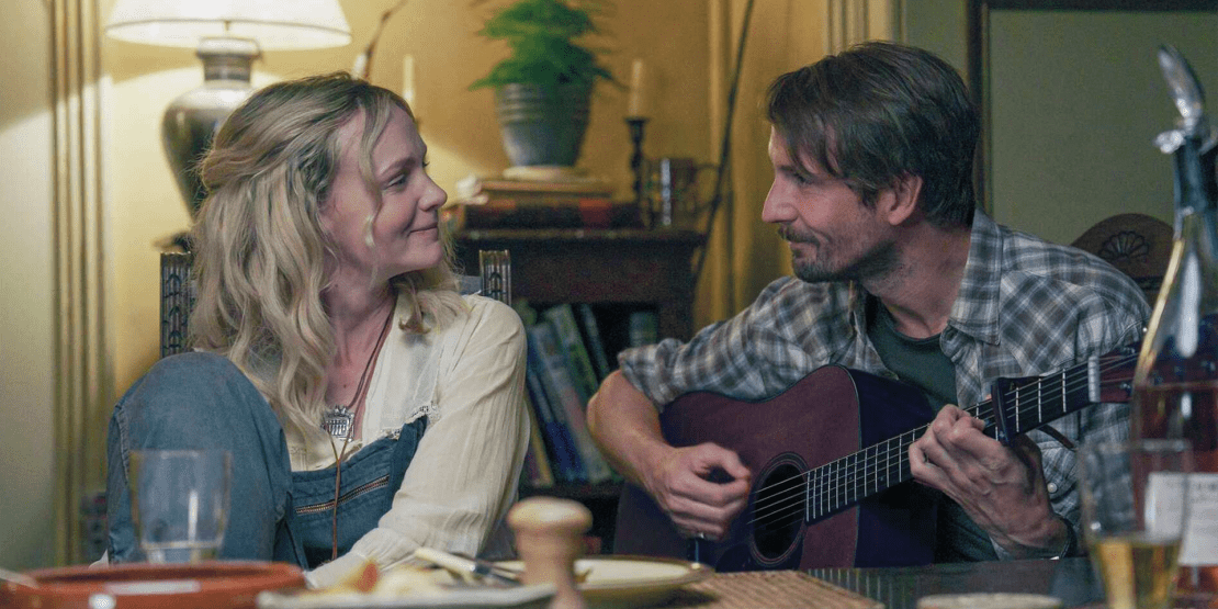 Woman smiles at a man playing acoustic guitar across a dining table during a scene from The Ballad of Wallis Island.