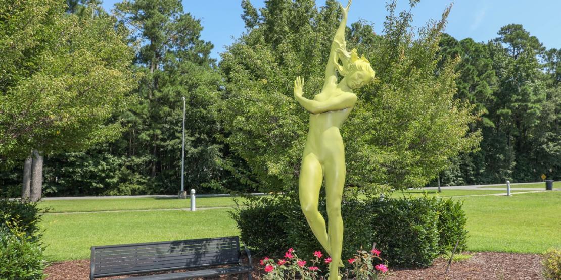 Bright green Crest of the Wave sculpture depicting a dancing figure in a landscaped garden on campus.
