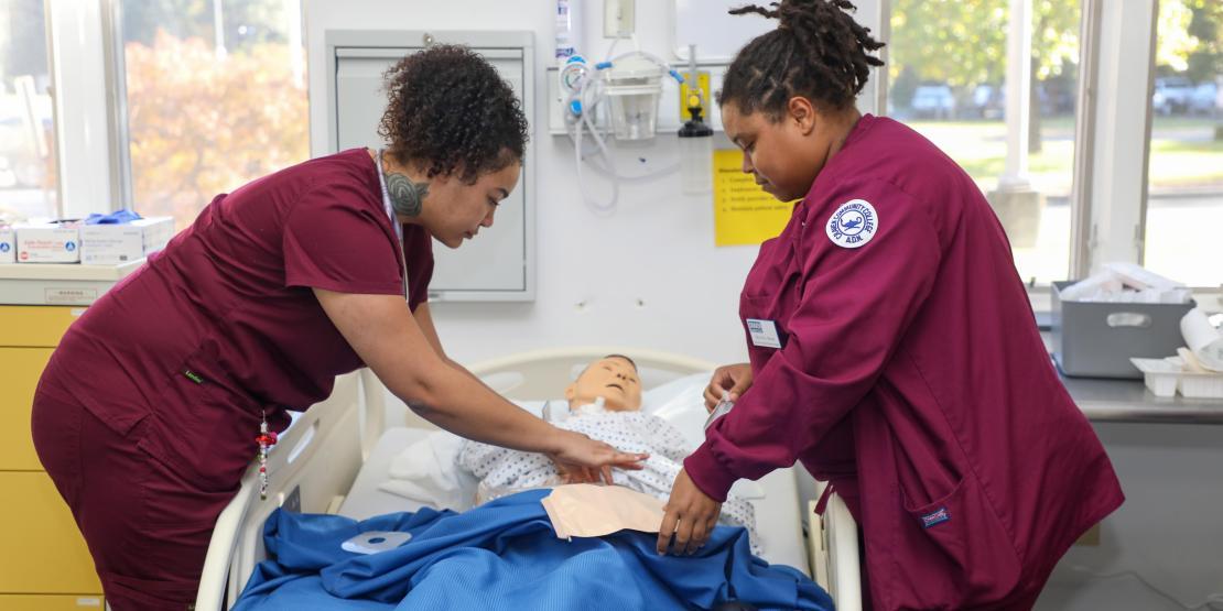 Nursing students in scrubs practice patient care using a medical training mannequin.