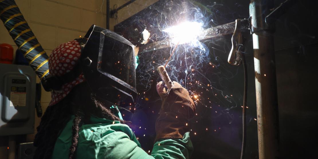 Welding student wearing protective gear welds a metal workpiece as sparks fly.