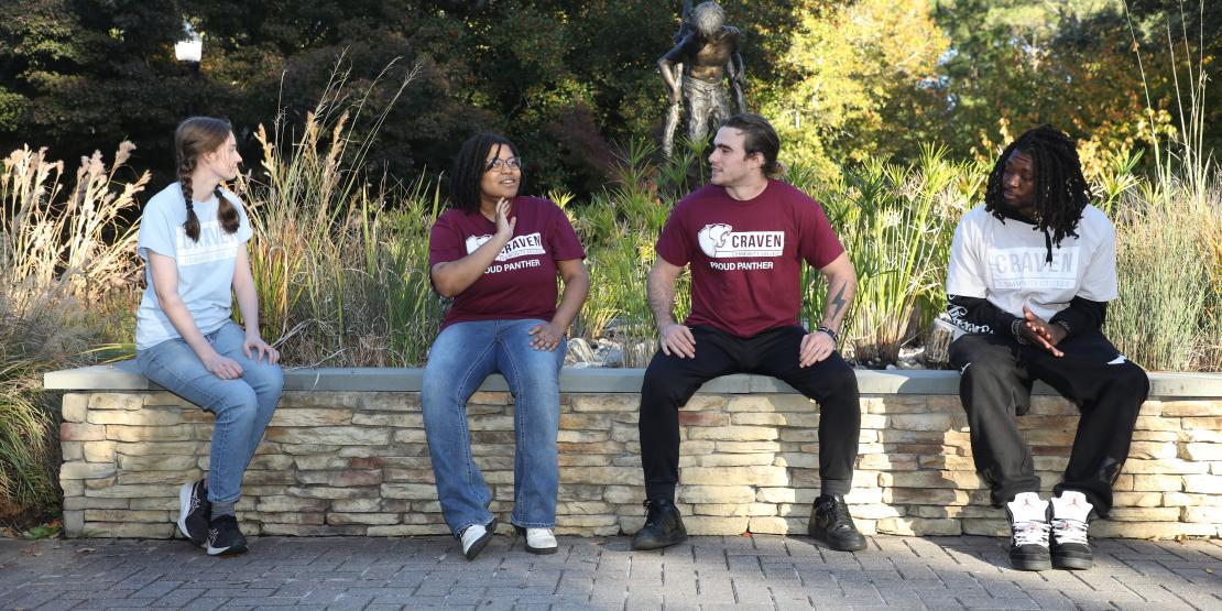 Four students sit spaced apart on a low stone wall in a landscaped campus courtyard, talking and relaxing outdoors.