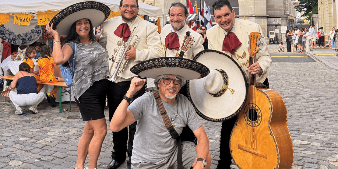 Mariachi musicians in traditional charro attire pose with violins, trumpets, and a large guitarrón while a smiling man in a sombrero kneels in front.