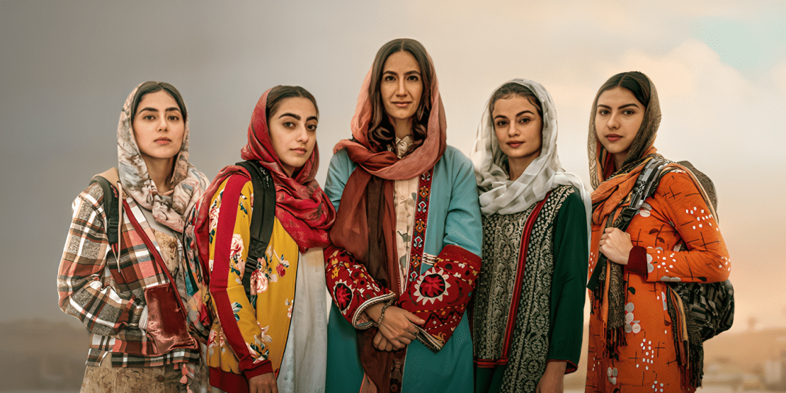 Five young women stand side by side in a dry, mountainous landscape, wearing colorful patterned dresses, shawls, and headscarves in shades of red, orange, green, and blue.