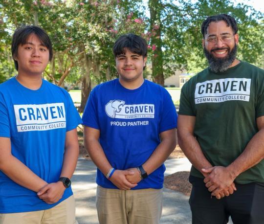 Three Craven Community College students wearing college T-shirts stand together outdoors on campus.