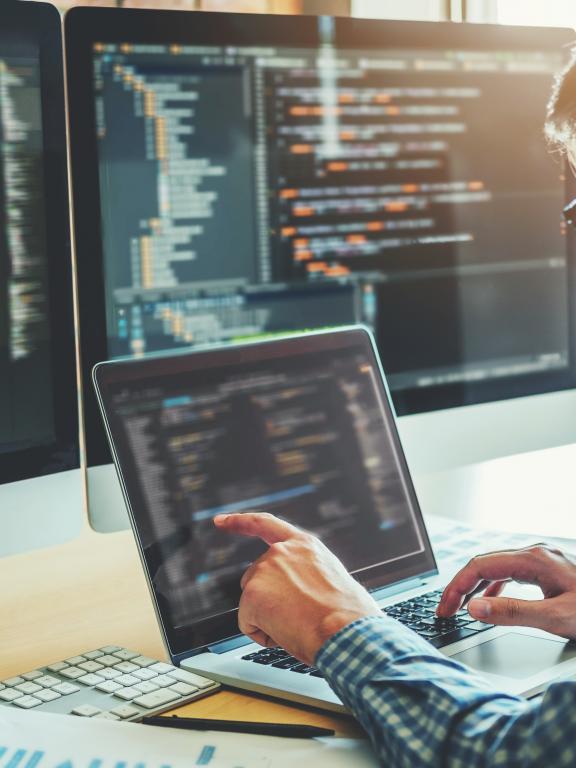 Male IT worker deciphers code on laptop in front of dual desktop monitors