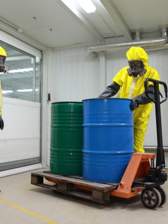 Two people in yellow hazmat suits transport a green and blue barrel of hazardous material on pull cart
