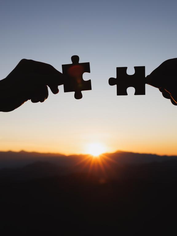 Silhouette of two hands holding up puzzle pieces in front of horizon during sunset