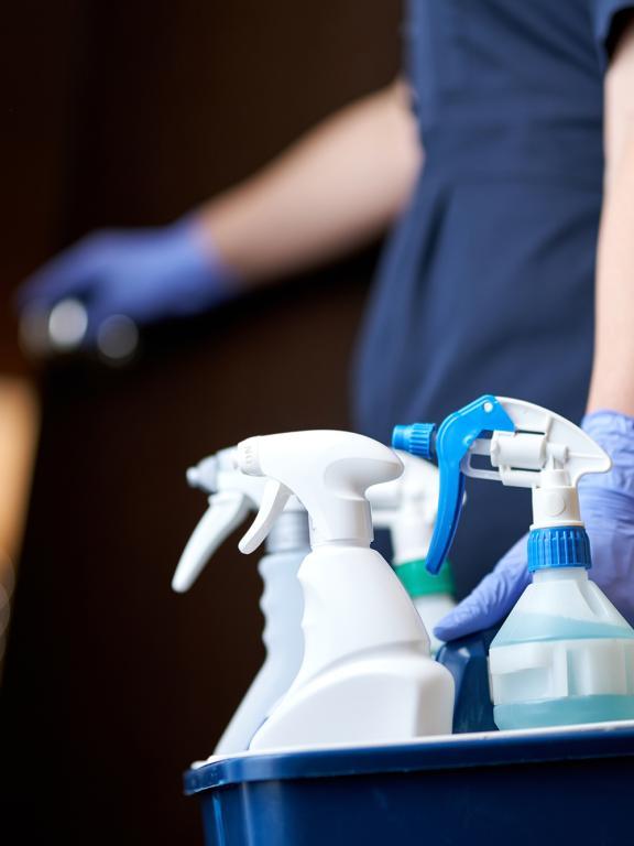 A cleaner wearing dark blue scrubs and gloves carries a bucket with spray bottles