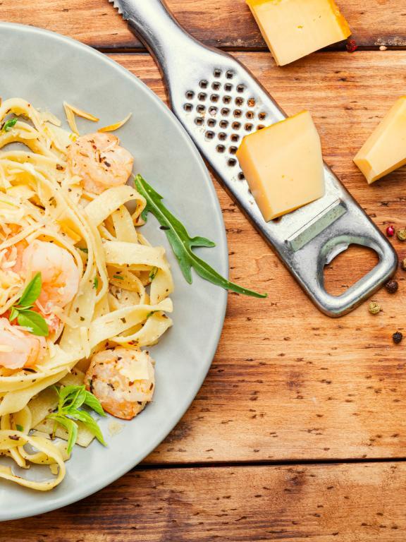 Plate of pasta and shrimp on wooden table next to grater and chunks of cheese and peppercorns