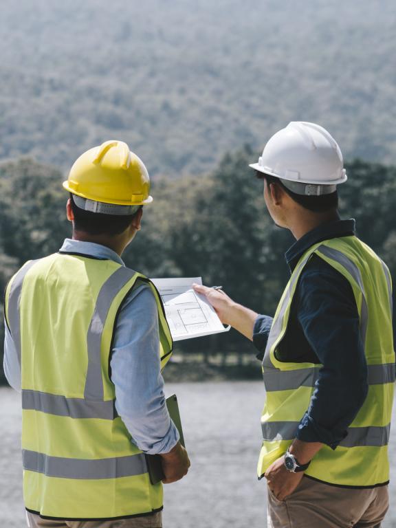 Two male forestry workers in hard hats and safety vests hold a blueprint and look out over a vast forest