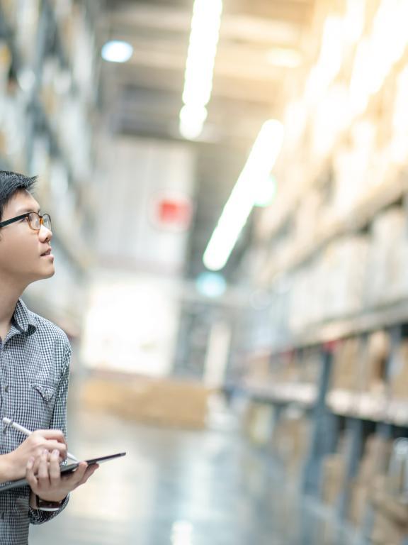 Young male worker taking stock of products in cardboard boxes on shelves in warehouse using digital tablet and pen