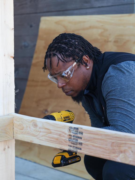 Construction student wearing safety glasses uses a drill to assemble a wooden frame.