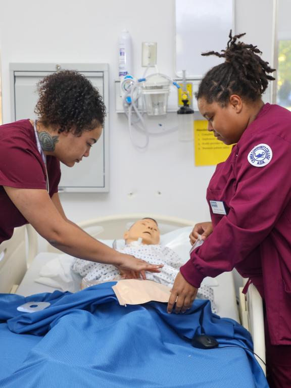 Nursing students in scrubs practice patient care using a medical training mannequin.