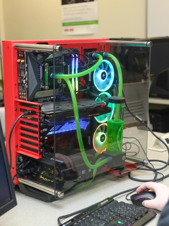 Information technology student works at a computer beside a custom-built PC with visible internal components and liquid cooling.