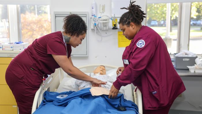 Nursing students in scrubs practice patient care using a medical training mannequin.