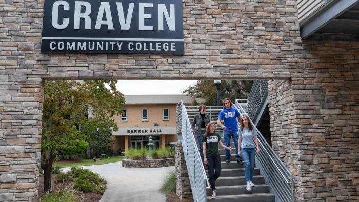 Craven Community College students walk down outdoor stairs beneath the campus entrance sign, with Barker Hall in the background.