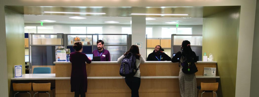 Three students stand at the counter while being helped by academic advisors under an illuminated sign reading "Admissions & Advising"