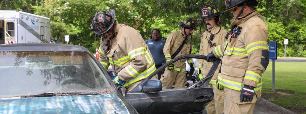Firefighting students in protective gear practice an emergency response around a vehicle.