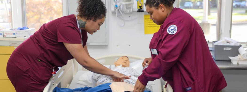 Nursing students in scrubs practice patient care using a medical training mannequin.