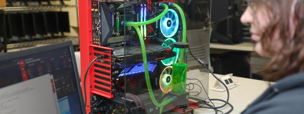 Information technology student works at a computer beside a custom-built PC with visible internal components and liquid cooling.