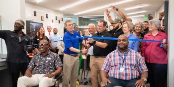 People gather in renovated barbershop as a blue ribbon is cut with oversized scissors 