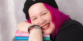 Craven alum Xena Casey smiles and leans over a pie of textbooks, wearing a black beret with silver star, black T-shirt, and bright pink hair
