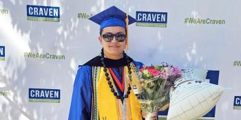 Craven CC alum Dominic Bradfield poses against Craven CC backdrop in cap and gown holding a bouquet of flowers and a balloon