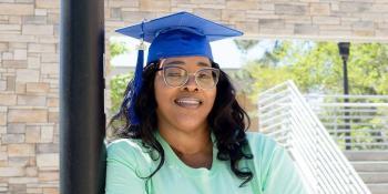 Craven alum Catrina Mack Cook crosses her arms and wears a grad cap in front of the Craven Community College sign