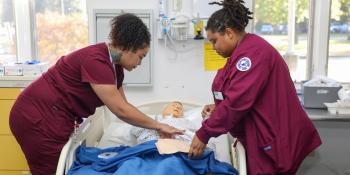 Nursing students in scrubs practice patient care using a medical training mannequin.